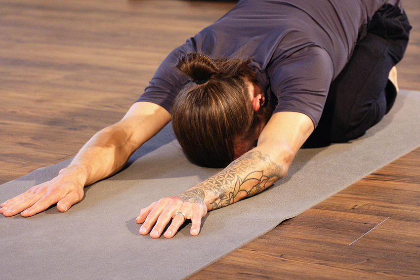 Der Yoga Lehrer zeigt ein leichtes Asana beim Yoga lernen. Er sitzt und das Gesicht liegt auf der Matte, die Hände liegen ausgestreckt vor ihm.