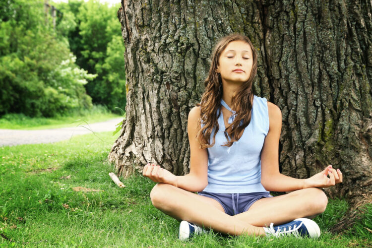 Teenager doing yoga and relaxing by siting in a grass in a park on a huge and old tree.
